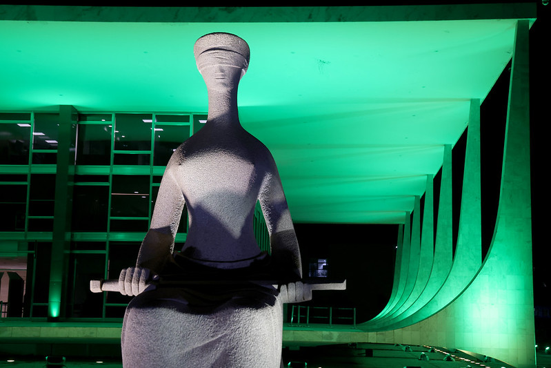 Imagem de estátua representando a Justiça em frente à fachada do STF iluminado à noite por luz verde
